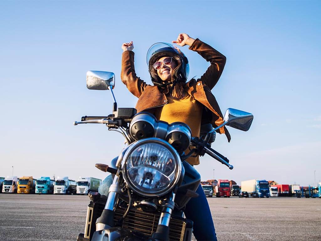 mulher-jovem-e-bonita-montada-em-moto-usando-oculos-escuros-com-capacete-e-com-as-maos-para-cima-usando-jaqueta-marron-e-blusa-amarela-com-caminhoes-estacionados-ao-fundo